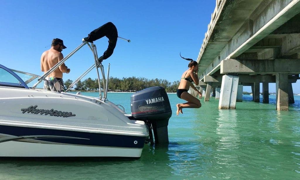 Person diving off a boat into turquoise water under a bridge on a sunny day.