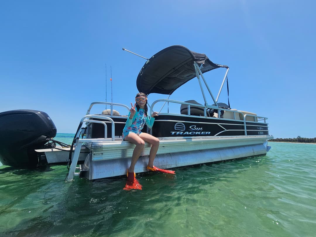 Child wearing snorkeling gear sitting on a Sun Tracker boat in clear water. Sunny day, outdoor fun.