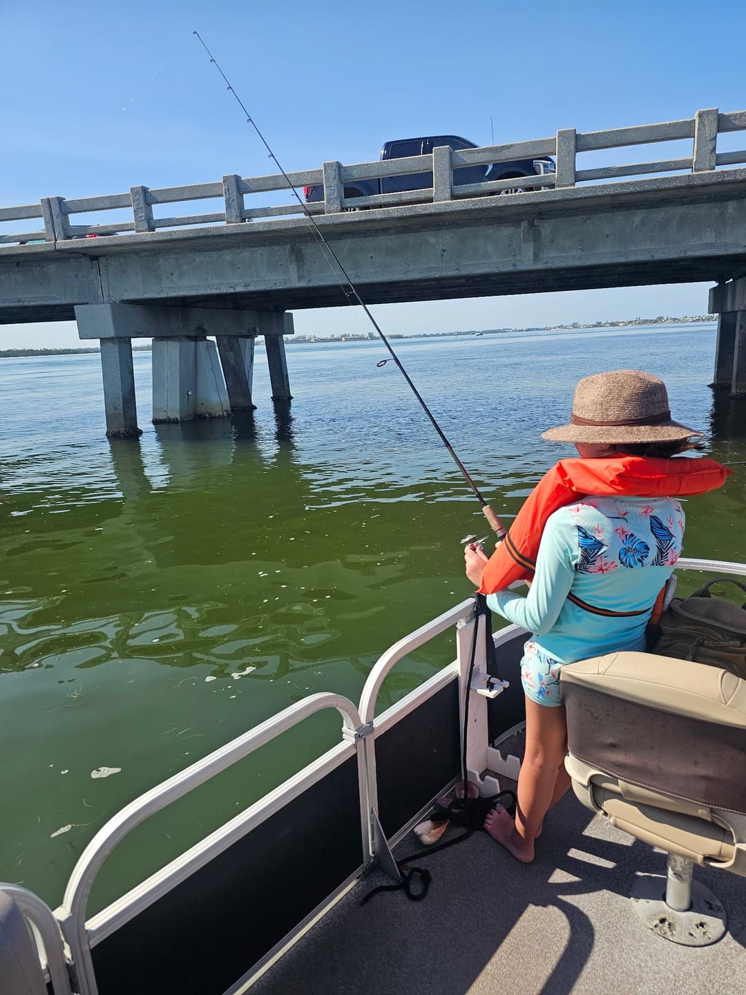 Child fishing from a boat under a bridge on a sunny day, wearing a sun hat and life jacket.